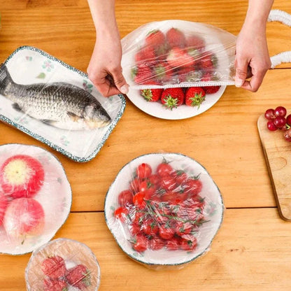 Hands using clear wrap to cover fruits and a fish on a wooden table.