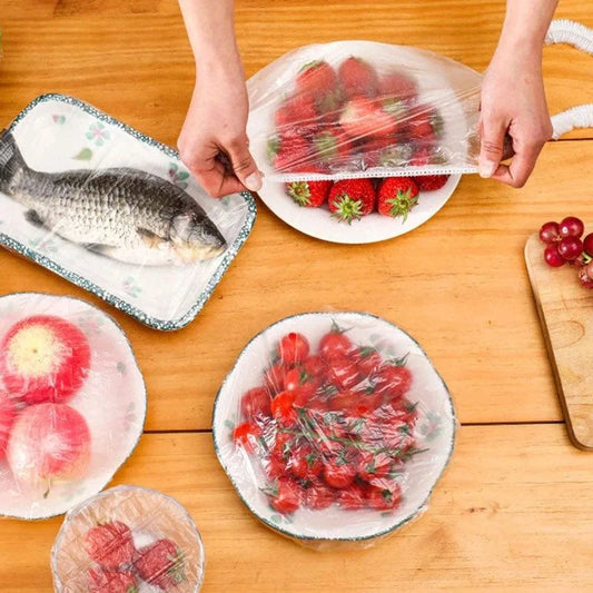 Hands using clear wrap to cover fruits and a fish on a wooden table.