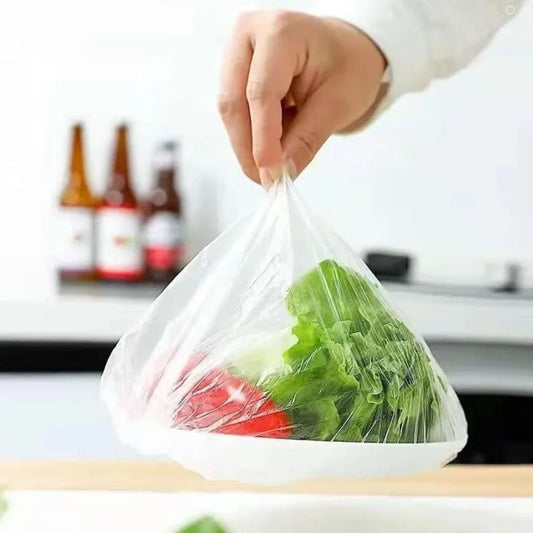 A hand holding a clear plastic bag filled with green lettuce and red tomato, with a blurred kitchen background and bottles in the distance.