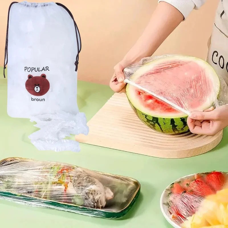 A clear stretch food cover being stretched over a watermelon on a wooden board, with a packaged meal covered in the same clear film on a tray and a white drawstring bag printed with a bear logo and the word 'broun' standing to the left. A person's hands and torso are visible in a kitchen-like setting.